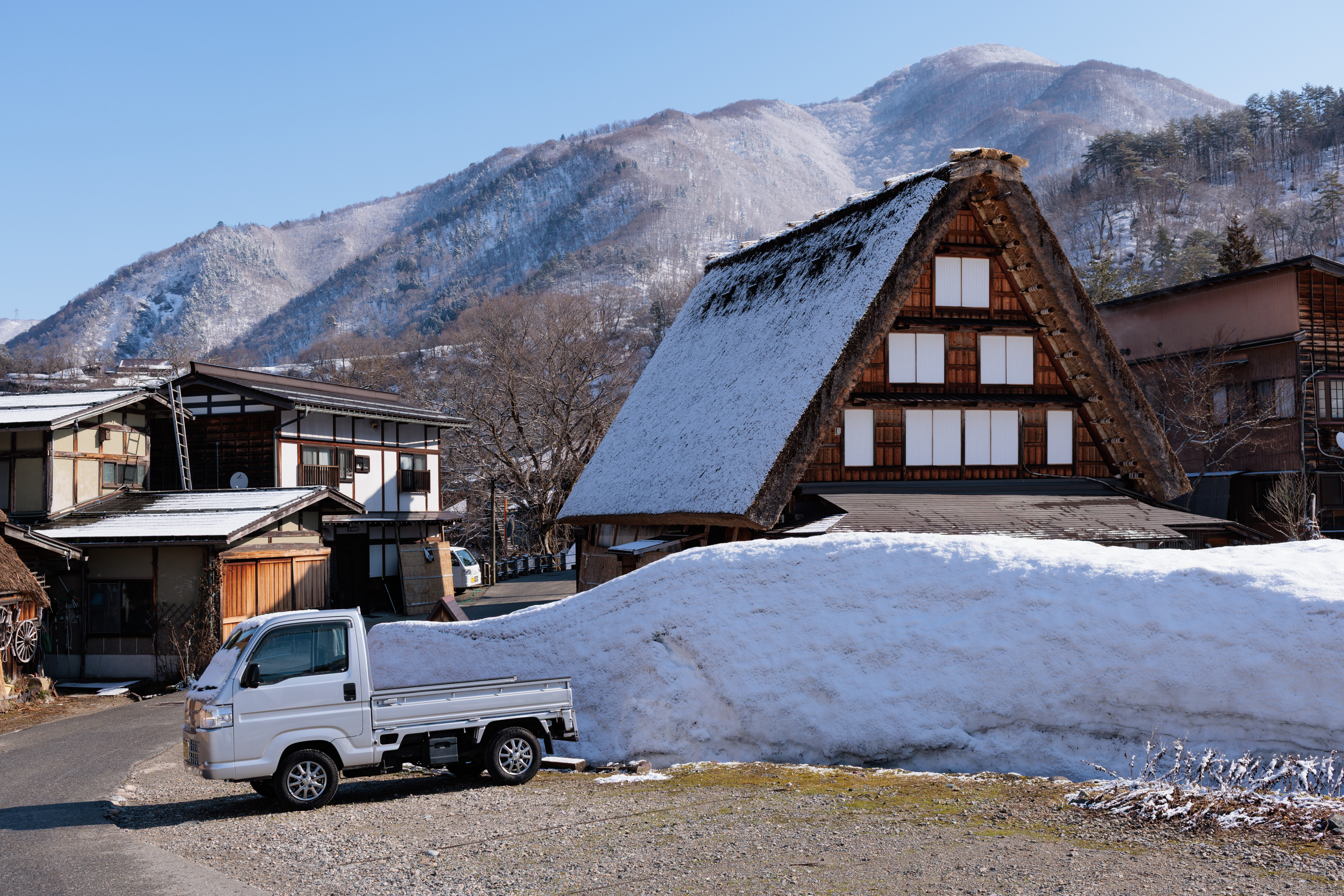A snowbank in front of a Gassho style home with a kei truck next to it